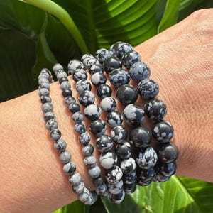 May include: A stack of six bracelets made of black and white snowflake obsidian beads. The bracelets are worn on a person's wrist.