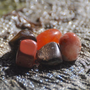 May include: A collection of five polished stones in shades of brown, red, and orange. The stones are arranged on a textured surface.