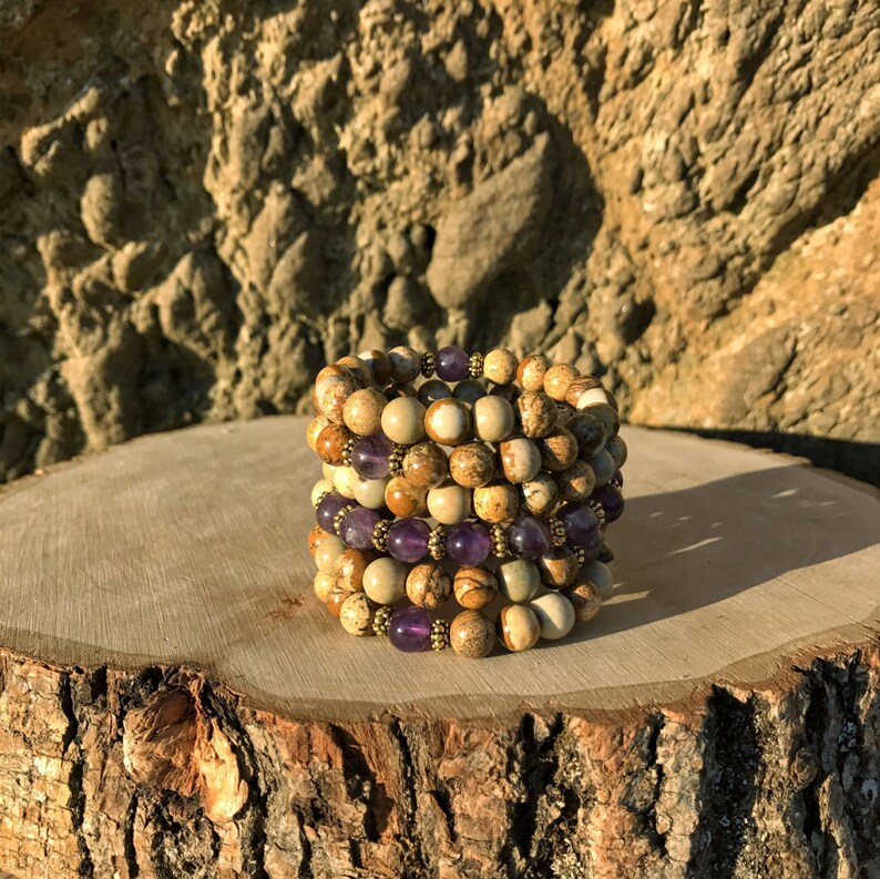 May include: A stack of four bracelets made with brown and white jasper beads and amethyst beads. The bracelets are stacked on a wooden surface.