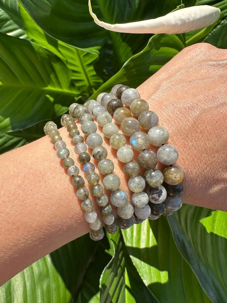 May include: A collection of beaded bracelets in shades of grey, brown, and green, with varying bead sizes, are stacked on a wrist. The bracelets feature polished, round beads. The background shows green leaves and a white flower.