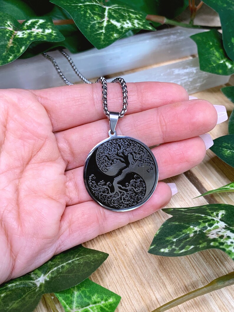 May include: A silver-toned necklace featuring a circular pendant with a black yin-yang design. The pendant showcases a detailed tree of life motif. The necklace is displayed against a backdrop of greenery and a wooden surface.