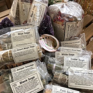 May include: An assortment of sage smudge sticks and Palo Santo wood in clear plastic bags. The sage bundles are labelled with their names, including White Sage, Black Sage, Blue Sage, Dragon's Blood Sage, Cedar Sage, Juniper Smudge, Desert Magic Sage, and White Sage & Lavender. The Palo Santo wood is labelled with the text "Palo Santo".