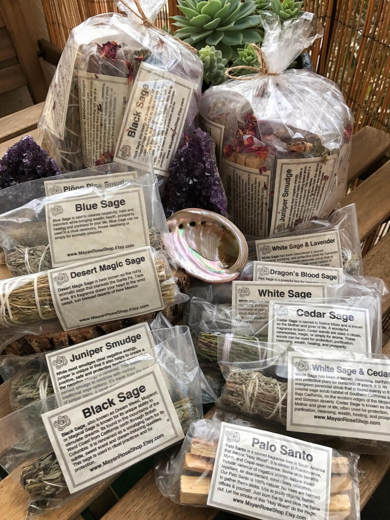 May include: An assortment of sage smudge sticks and Palo Santo wood in clear plastic bags. The sage bundles are labelled with their names, including White Sage, Black Sage, Blue Sage, Dragon's Blood Sage, Cedar Sage, Juniper Smudge, Desert Magic Sage, and White Sage & Lavender. The Palo Santo wood is labelled with the text "Palo Santo".