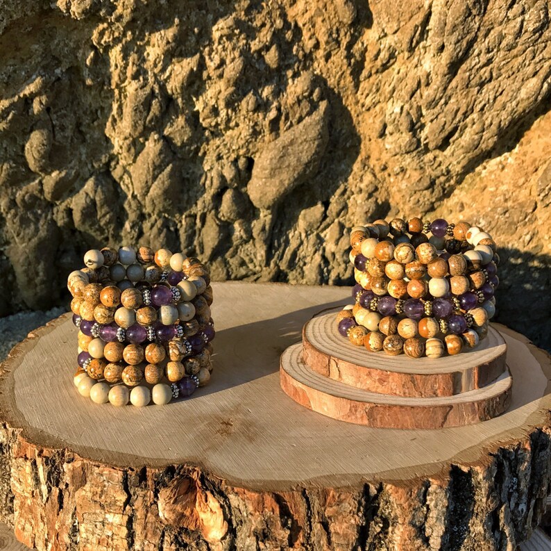 May include: A set of stacked bracelets made of brown and white jasper beads with a row of purple amethyst beads in the center. The bracelets are displayed on a wooden surface.