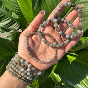 May include: A collection of labradorite beaded bracelets in shades of grey and green. The bracelets, of different sizes, are displayed on a hand, with a stack on the wrist. The background shows large green leaves.