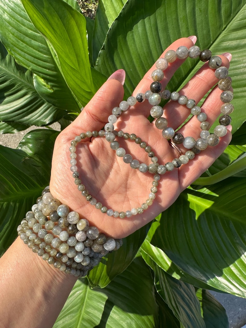 May include: A collection of labradorite beaded bracelets in shades of grey and green. The bracelets, of different sizes, are displayed on a hand, with a stack on the wrist. The background shows large green leaves.