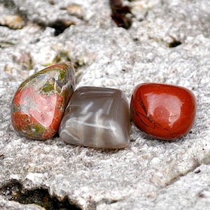 Puede incluir: Tres piedras lisas y pulidas en fila sobre una superficie gris texturizada. Las piedras son rojas con marcas verdes y blancas, grises con vetas blancas y rojas con un acabado brillante.