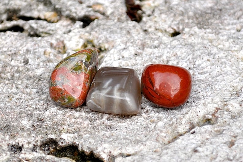 Puede incluir: Tres piedras lisas y pulidas en fila sobre una superficie gris texturizada. Las piedras son rojas con marcas verdes y blancas, grises con vetas blancas y rojas con un acabado brillante.