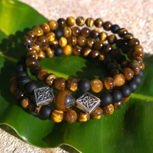 Puede incluir: Un brazalete de triple vuelta hecho con cuentas de ojo de tigre y cuentas negras. El brazalete tiene dos abalorios plateados con un diseño de nudo celta.