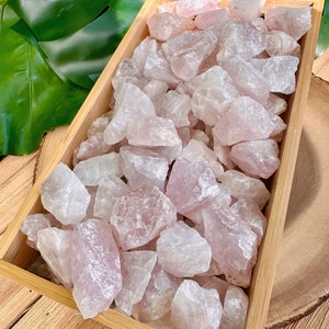 May include: A wooden tray filled with raw rose quartz crystals. The crystals vary in size and shape, displaying shades of pale pink and translucent white. The tray is made of light-colored wood, and the background includes green leaves and a wooden surface.