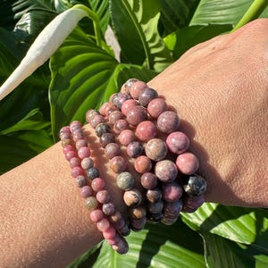 May include: A stack of pink and grey gemstone bracelets. The bracelets are made of round beads and are stacked on a person's wrist.