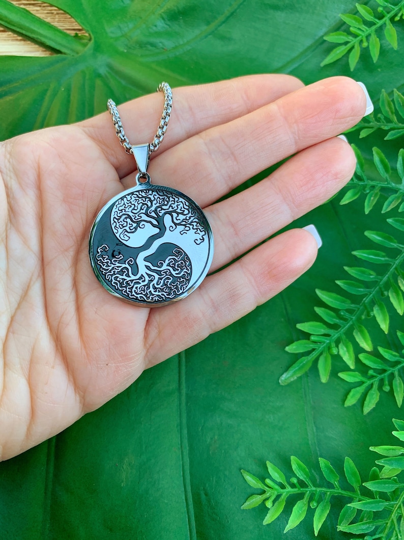 May include: A silver-toned pendant necklace featuring a Yin and Yang design with a tree of life motif. The pendant is circular and hangs from a matching chain. The necklace is displayed against a green background.