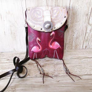 May include: A burgundy leather crossbody bag with a flap closure. The flap is cream-colored with embroidered swirls and a silver buckle. Two pink flamingos are appliqued on the front. The bag has a brown leather strap and decorative tassels.