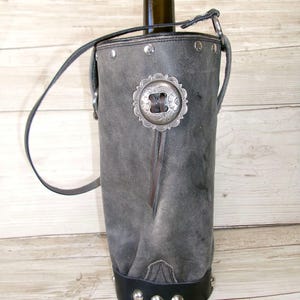 May include: A cylindrical gray leather wine bottle carrier featuring a black leather base with silver metal studs. The carrier includes a thin black leather shoulder strap and a decorative silver-toned metal concho ornament centered on the front.