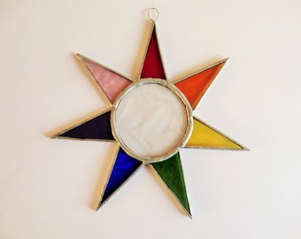 Könnte beinhalten: Ein farbenfroher Glasstern mit einer zentralen kreisförmigen Öffnung. Der Stern hat acht Spitzen in verschiedenen Farben, darunter Rot, Orange, Gelb, Grün, Blau und Rosa, mit einem silbernen Metallrahmen. Oben befindet sich eine kleine Metallschlaufe zum Aufhängen.