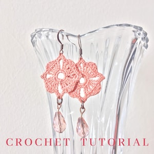May include: A pair of crocheted flower earrings with crystal drops. The earrings are a light pink color and are hanging from a glass vase.