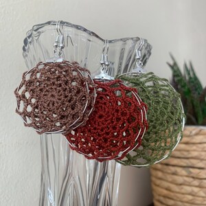 May include: Three handmade crochet earrings in shades of brown, red, and green. Each earring features a circular design with a delicate, open weave pattern, suspended from silver-colored hooks. The earrings are displayed in a clear glass vase.