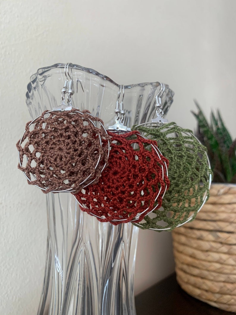 May include: Three handmade crochet earrings in shades of brown, red, and green. Each earring features a circular design with a delicate, open weave pattern, suspended from silver-colored hooks. The earrings are displayed in a clear glass vase.