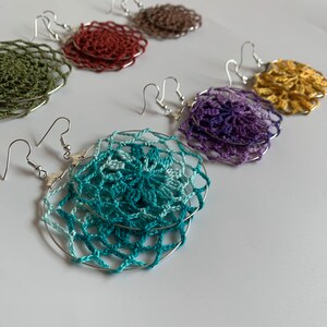 May include: A collection of handmade crochet earrings in various colors, including teal, purple, gold, and olive green. Each earring features a delicate, circular design with a silver-tone hoop and hook closure. The earrings are displayed on a white surface.