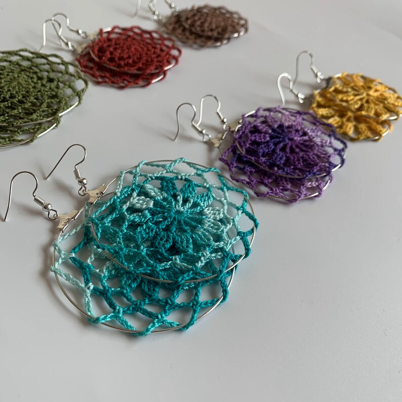 May include: A collection of handmade crochet earrings in various colors, including teal, purple, gold, and olive green. Each earring features a delicate, circular design with a silver-tone hoop and hook closure. The earrings are displayed on a white surface.