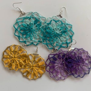 May include: Handmade crochet earrings in teal, gold, and purple. Each pair features a circular design with a floral pattern, suspended from silver-tone ear wires. The earrings are displayed on a white surface, showcasing the intricate details of the crochet work.