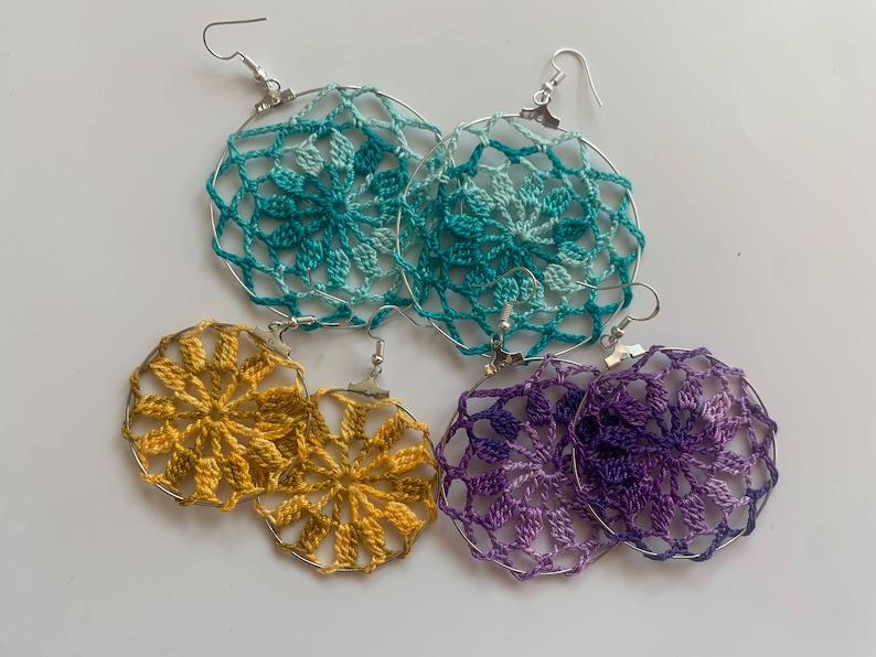 May include: Handmade crochet earrings in teal, gold, and purple. Each pair features a circular design with a floral pattern, suspended from silver-tone ear wires. The earrings are displayed on a white surface, showcasing the intricate details of the crochet work.