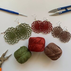 May include: Handmade crochet earrings in olive green, rust, and brown, with silver-tone hoops. The image also shows spools of matching yarn, a crochet hook, scissors, and pliers, all arranged on a white surface.