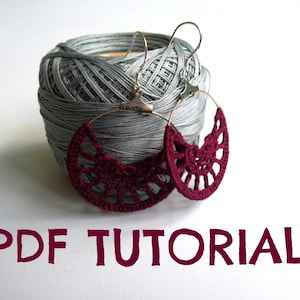 Può includere: Un paio di orecchini a uncinetto color borgogna con cerchietti d'argento. Gli orecchini sono esposti su un gomitolo di filato grigio. Il testo "PDF TUTORIAL" è scritto in lettere color borgogna sotto gli orecchini.