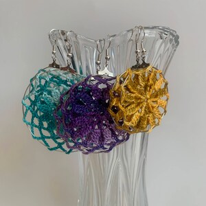 May include: Three handmade crochet earrings in teal, purple, and gold, each with a circular design and silver-colored ear wires. The earrings are displayed in a clear glass vase against a white background. The crochet work has a delicate, open weave.