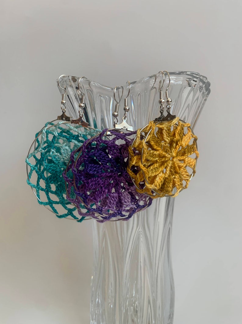 May include: Three handmade crochet earrings in teal, purple, and gold, each with a circular design and silver-colored ear wires. The earrings are displayed in a clear glass vase against a white background. The crochet work has a delicate, open weave.