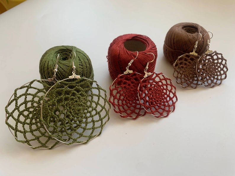 May include: Handmade crochet earrings in olive green, burgundy, and brown, with silver-tone hoops. The earrings are displayed with matching yarn balls, showcasing the craft's materials and colors. A unique accessory for any outfit.