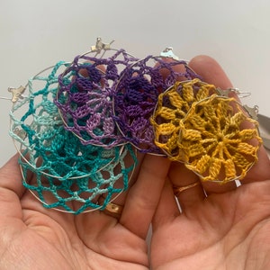 May include: A collection of handmade crochet earrings in various colors, including teal, purple, and gold. The earrings feature intricate floral patterns and are attached to silver hoops. A craft item.