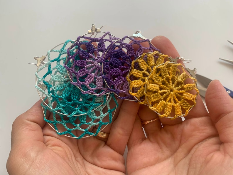 May include: A collection of handmade crochet earrings in various colors, including teal, purple, and gold. The earrings feature intricate floral patterns and are attached to silver hoops. A craft item.