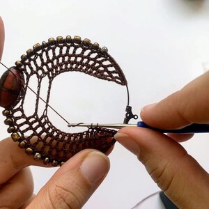 以下が含まれることがあります： 茶色のビーズと金属製の留め具が付いた、茶色のクロシェ編みフープのクローズアップ。人がクロシェフックと青い糸を使ってフープを持っています。
