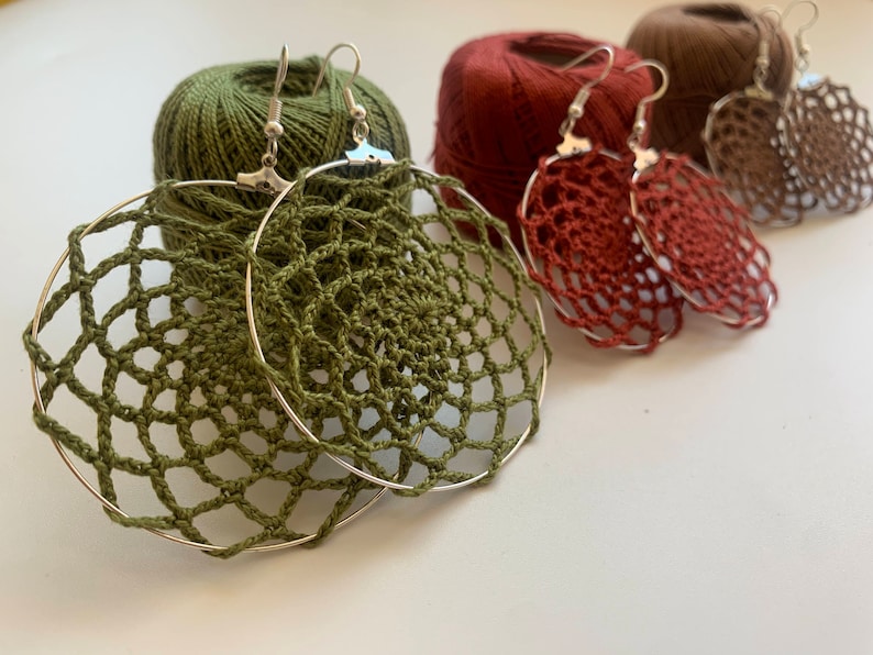 May include: Handmade crochet earrings in olive green, rust, and brown. The earrings feature a delicate, openwork design and are suspended from silver-tone hoops. The image also shows spools of matching yarn, suggesting a craft or DIY theme.