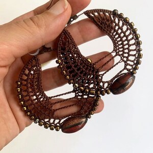 以下が含まれることがあります： ゴールドビーズと赤褐色の石のアクセントが付いた、茶色のクロシェ編みフープイヤリングのペアです。