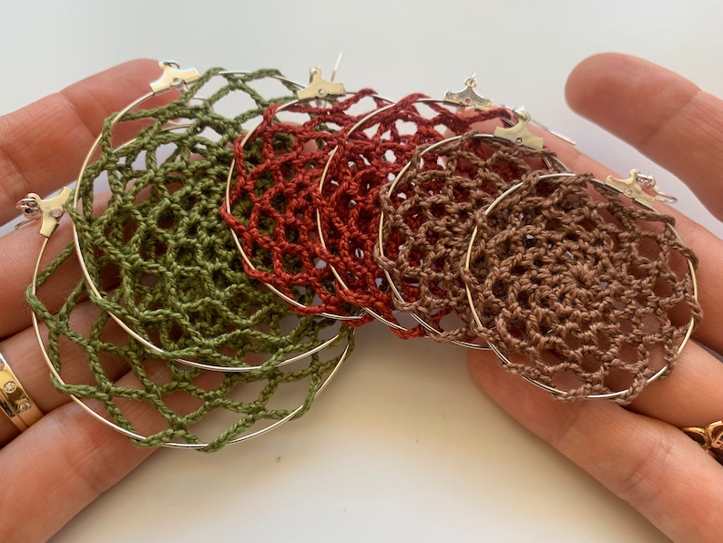 May include: A collection of handmade hoop earrings in shades of green, rust, and brown. Each earring features a crocheted web design within a silver-tone hoop. The earrings are displayed in a person's hands.