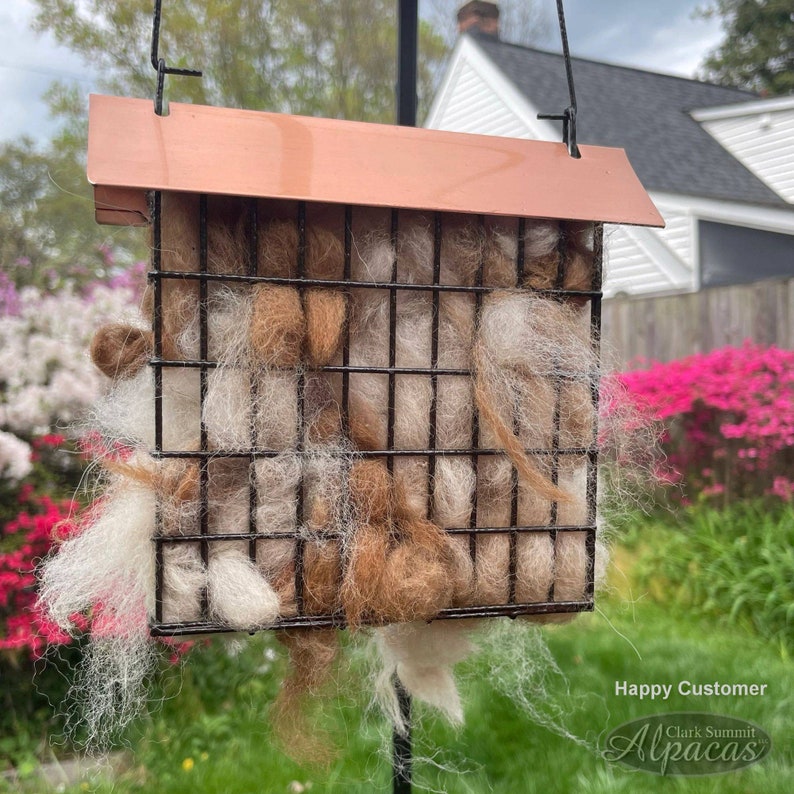 May include: A bird feeder filled with alpaca fleece in shades of white and brown. The feeder is made of metal with a copper-colored roof. The feeder is hanging from a black wire. The text "Happy Customer" and "Clark Summit Alpacas" is visible in the image.