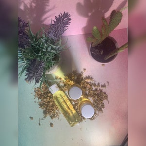 May include: A set of two small yellow jars with white lids and a clear glass bottle with a yellow liquid. The jars and bottle are on a white surface with dried yellow flowers.