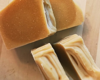 May include: Four bars of handmade soap, two are a light brown color and two are a lighter beige color with swirls. The soap bars are on a wooden surface.