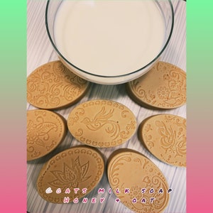 Puede incluir: Seis barras de jabón de leche de cabra de forma ovalada con varios diseños decorativos. El jabón es de color marrón claro y los diseños son de color marrón oscuro. El texto "OATS MILK SOAP HONEY + OAT" está impreso en una de las barras.