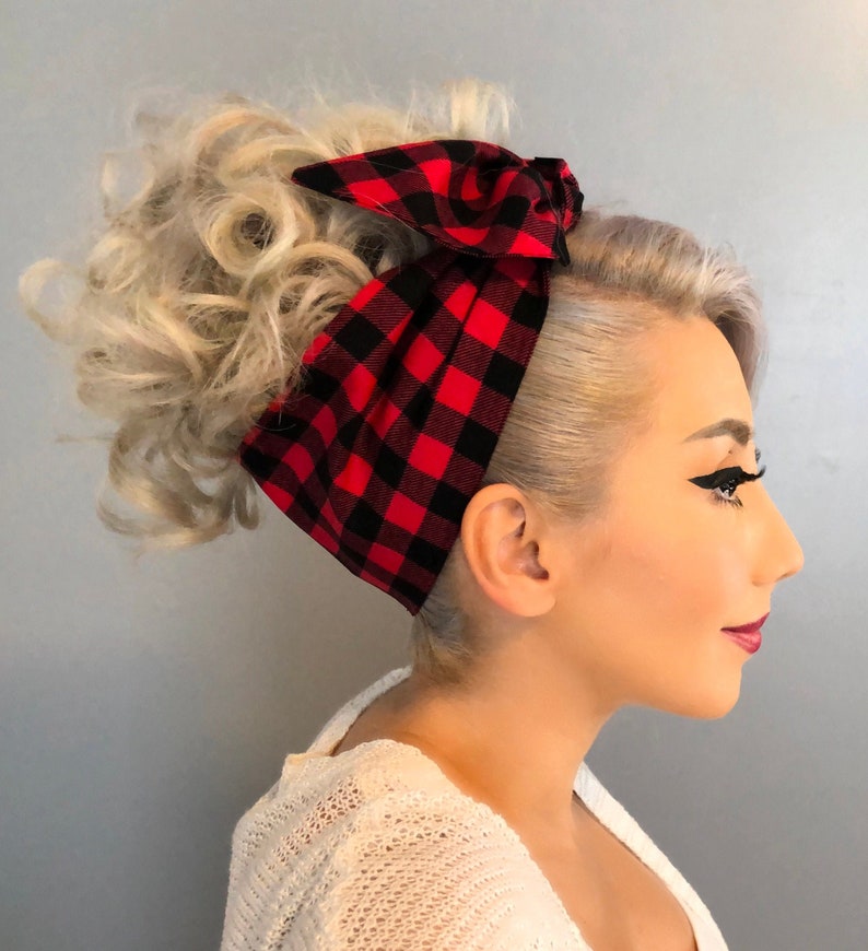 May include: A red and black plaid fabric headband tied in a bow. The headband is wrapped around a hairstyle with blonde curls. The person is wearing a white sweater.