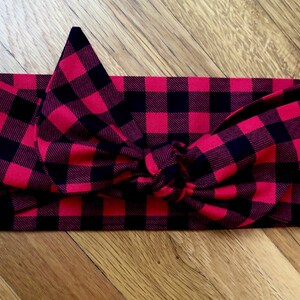 May include: A red and black buffalo plaid headband with a knotted bow. The fabric is a classic plaid pattern, with large, evenly spaced squares. The headband is designed to be worn around the head.