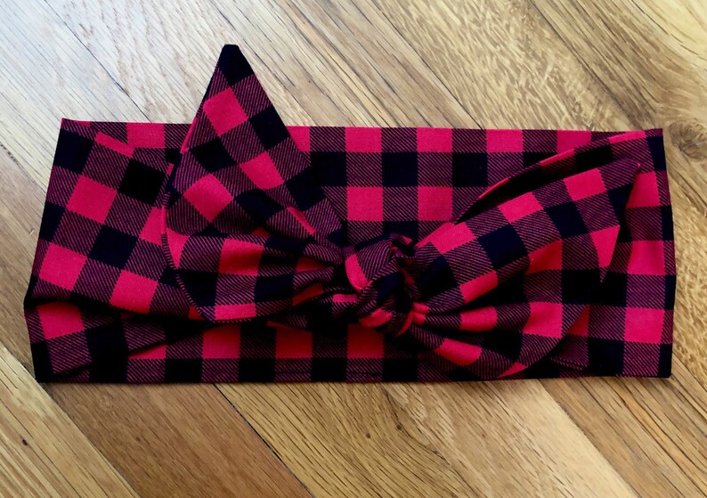 May include: A red and black buffalo plaid headband with a knotted bow. The fabric is a classic plaid pattern, with large, evenly spaced squares. The headband is designed to be worn around the head.
