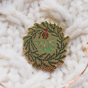 Op de afbeelding: Een goudkleurige emaille pin in de vorm van een kerstkrans met groene bladeren, een rode strik en maretak. De pin ligt op een witte, getextureerde gebreide achtergrond, met koperen lichtslingers.