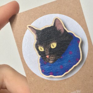 Op de afbeelding: Een houten pin met een zwarte kat die een blauwe en paarse sjaal draagt. De pin is op een bruine kaart met de tekst "Wooden pin by: the illustrationist illustrationist.etsy.com".