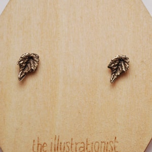 以下が含まれることがあります： 木製のディスプレイカードに置かれた、金色の葉の形をしたスタッドピアスのペア。カードには「the illustrationist」と「illustrationist.etsy.com」の文字が書かれています。