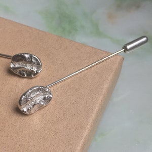 May include: Two silver-colored lapel pins, each featuring a coffee bean design. The pins are on a brown surface with the word "Illustrator" partially visible. The background has a green and white marbled pattern.