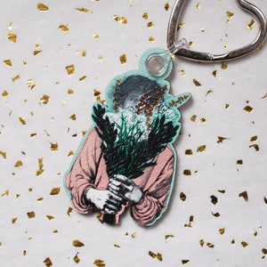 May include: A pink and green wooden keychain featuring a person holding a bouquet of flowers. The keychain has a silver heart-shaped ring.
