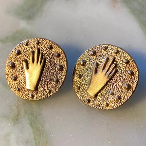 May include: A pair of gold-toned earrings with a raised hand design on each earring. The earrings are round and have a textured surface.